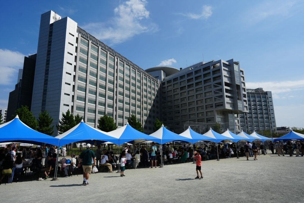 屋外の広場に複数のテントが並び、大勢の来場者が歩いたり列に並んだりしている様子